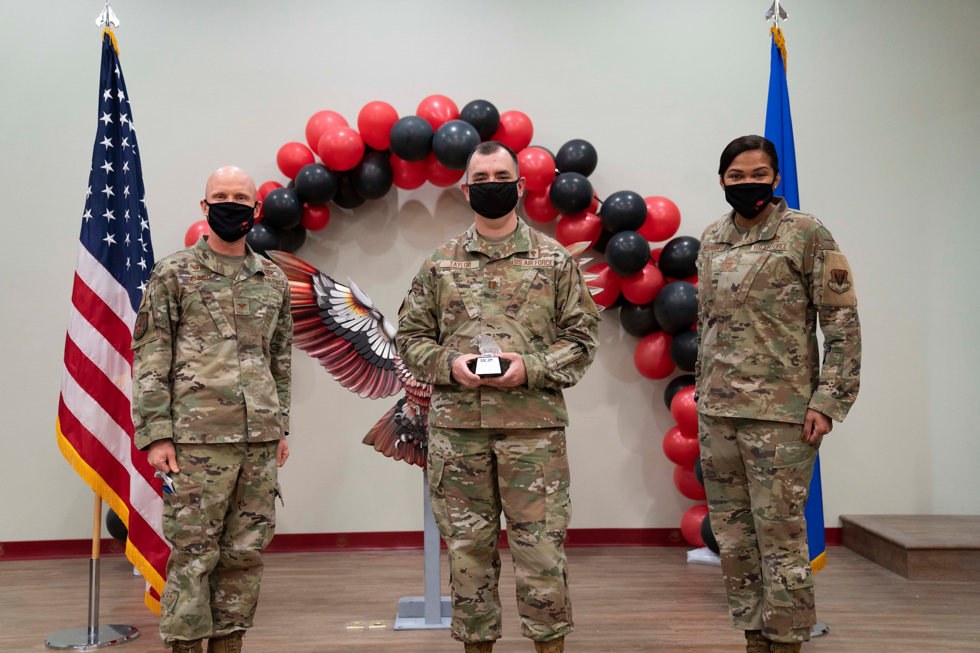 Capt. James Taylor stands in between Col. Eric C. Schmidt and Chief Master Sgt. Adrienne Warren as he holds his award at stomach level. the award is an shape of an American eagle head, facing left, made of glass with a wood plaque at the bottom with the award's name, his name and a "Thank you for your ministry!"