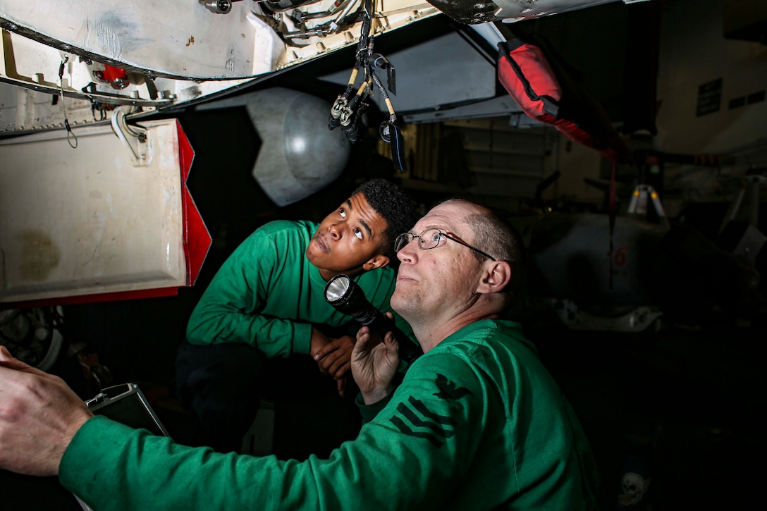Two men inspect the underside of an aircraft. One has a flashlight.