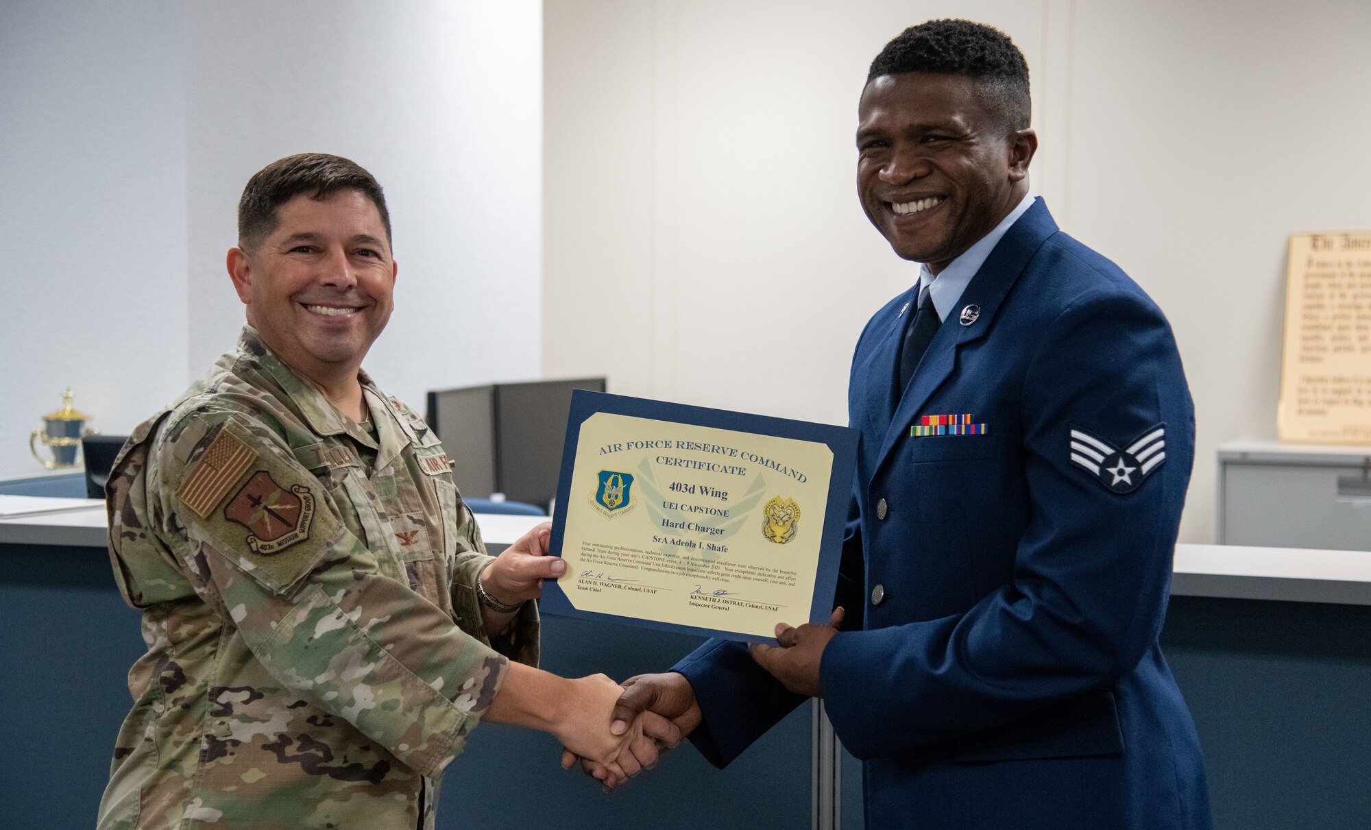 Col. Trujillo left, and Senior Airman Shafe, right, hold a certificate together and shake hands
