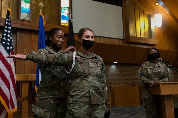 An airman stands with her arm raised as another pins the white rope on her uniform.