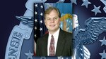 Head and shoulders shot of a white man in a dark suit in front of the US and DLA flags.