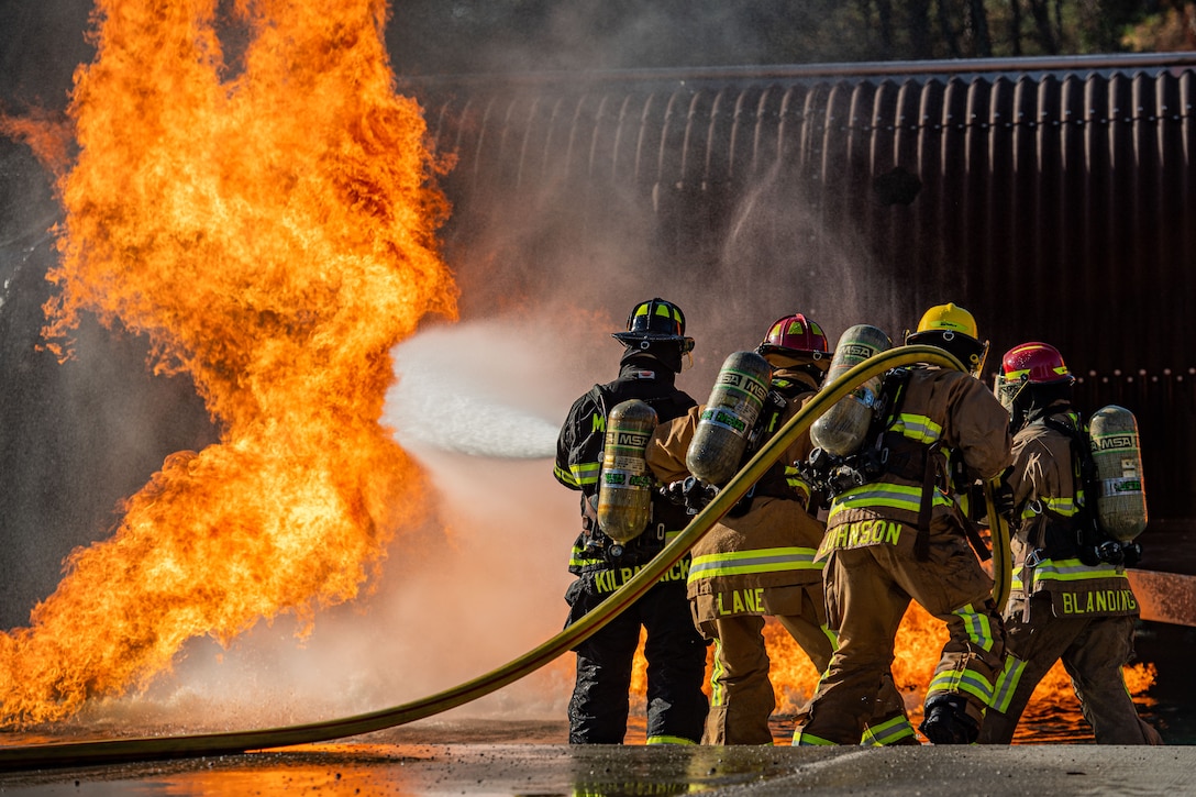 Airmen in firefighting gear use a hose to put out a fire.