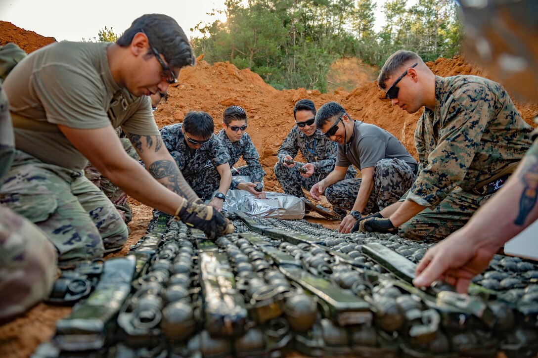 U.S. and Japanese troops work on grenades while kneeling in the dirt.