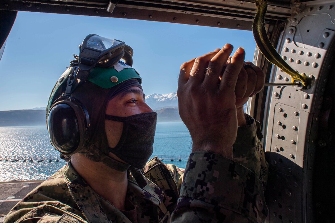 A sailor wearing a face mask uses a screwdriver to tighten a helicopter’s handle aboard a ship at sea.