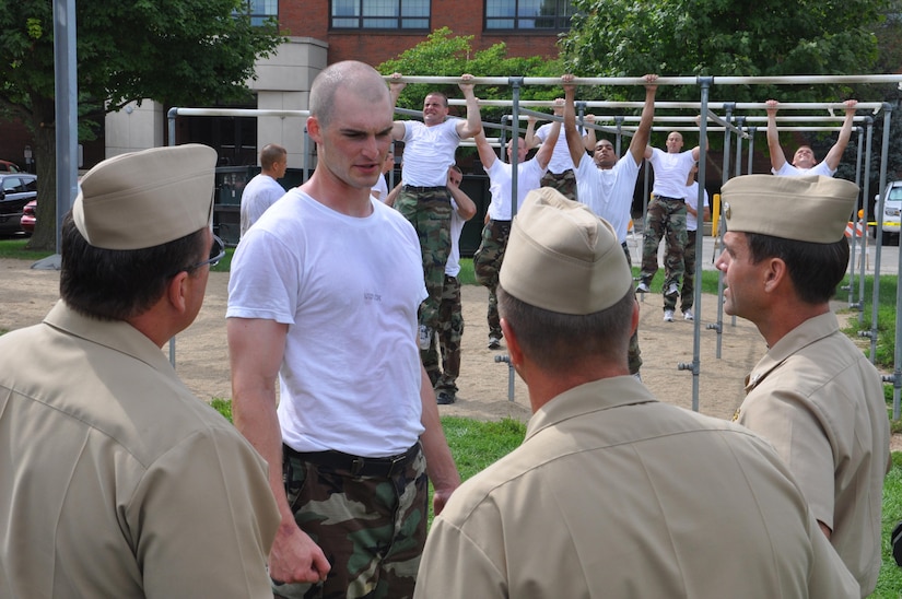 Sailors do pull ups in the background as a recruit speaks to three men in khaki uniform.