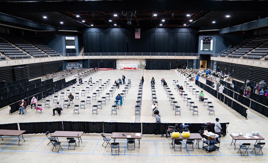 Wide view of large gym-type room with rows of chairs, some with people sitting in them.