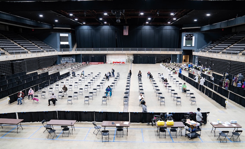 Wide view of large gym-type room with rows of chairs, some with people sitting in them.