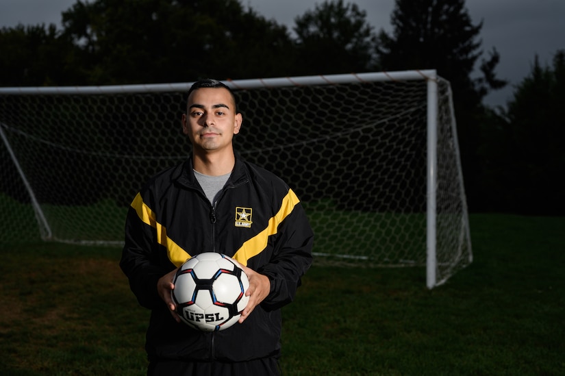 A soldier in an Army tracksuit holds a soccer ball in front of a goal.