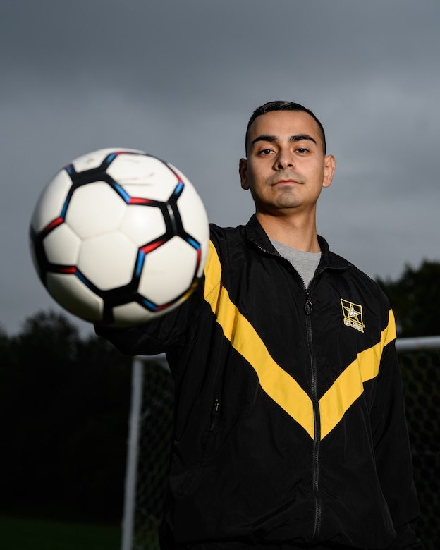 A soldier in an Army tracksuit holds up a soccer ball.