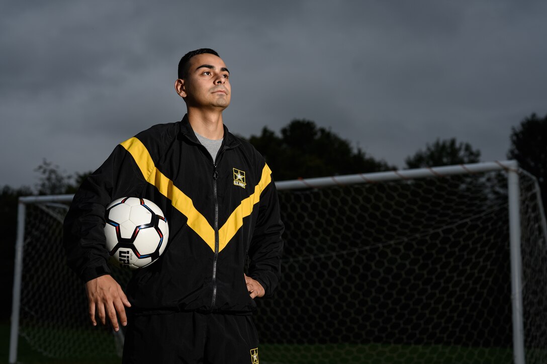 A soldier in an Army track suit poses with a soccer ball in front of a goal.