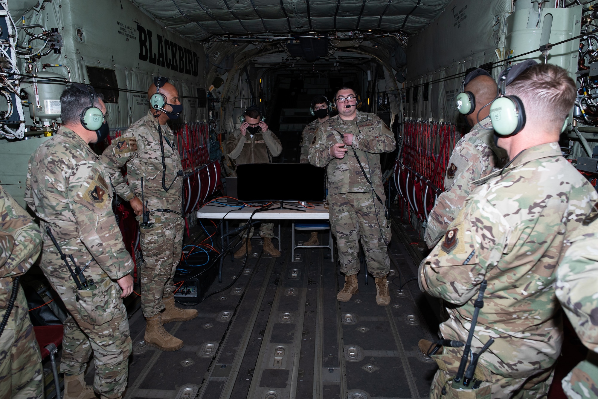 Airmen wearing headsets stand in a military cargo aircraft listening to another Airman standing in front of a table with computers on it.
