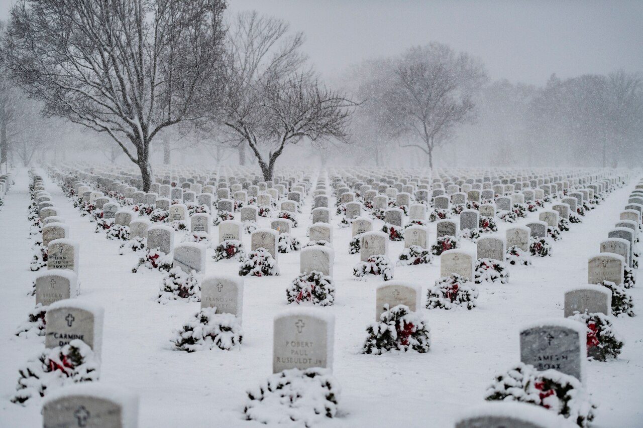 Snow falls in a cemetery covering headstones and wreaths.