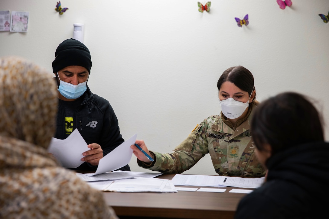 A female soldier wearing a face mask and a volunteer sit at a desk checking Afghan evacuees into a COVID-19 vaccination clinic.