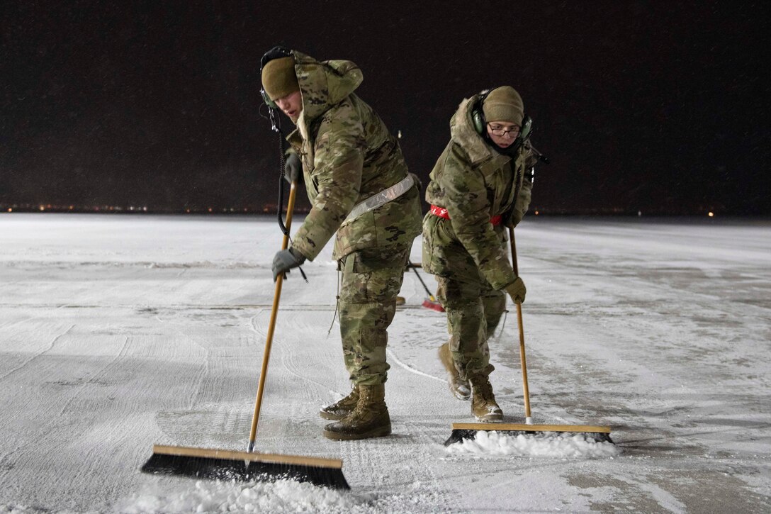 Two airmen dressed in winter gear push brooms to remove snow from a tarmac.