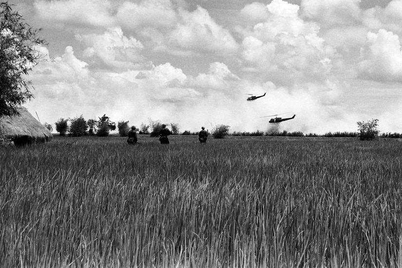 Soldiers walk through high grass while two helicopters patrol overhead.
