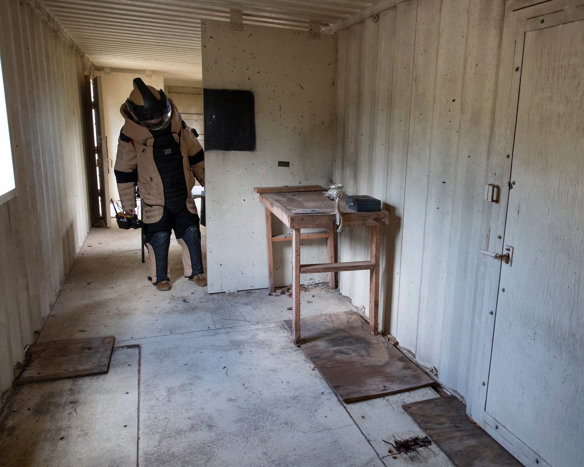 An EOD tech, wearing a bomb suit, peers into a room at an IED device during an IED training scenario.