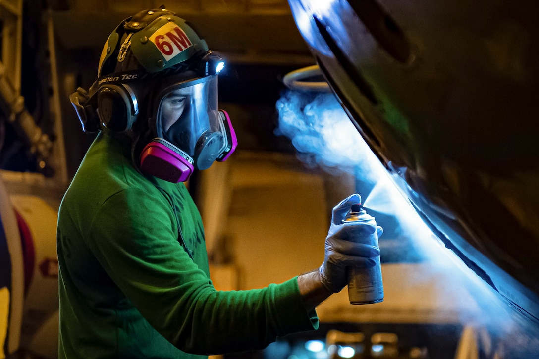 A sailor wearing a face shield spray-paints an aircraft.