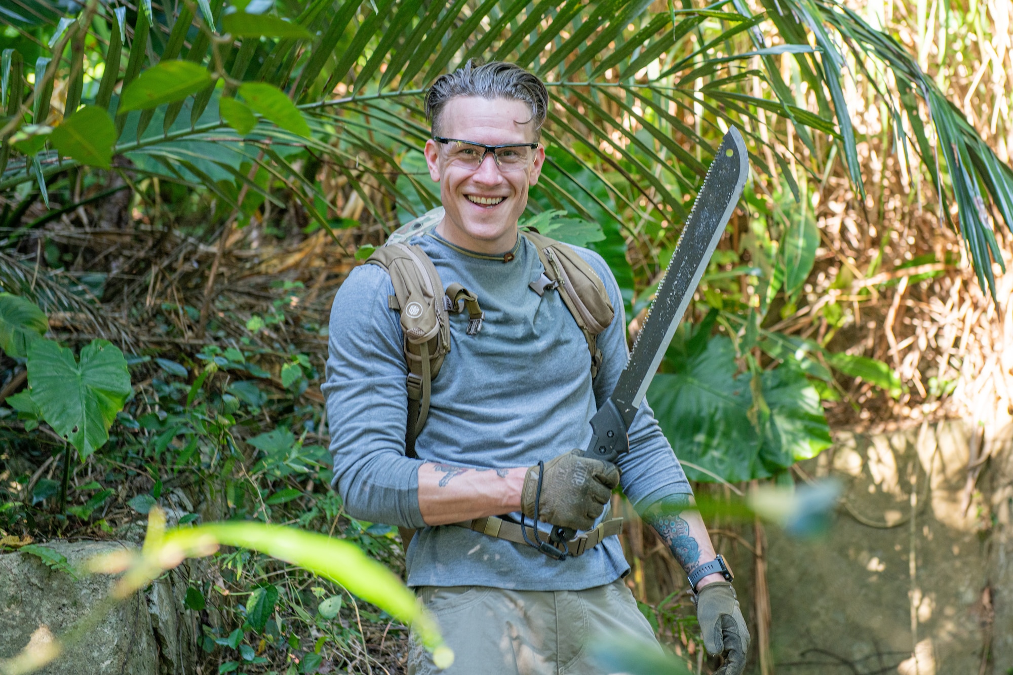 An Airman stands poses in the middle of the vegetation that he is clearing with a machete