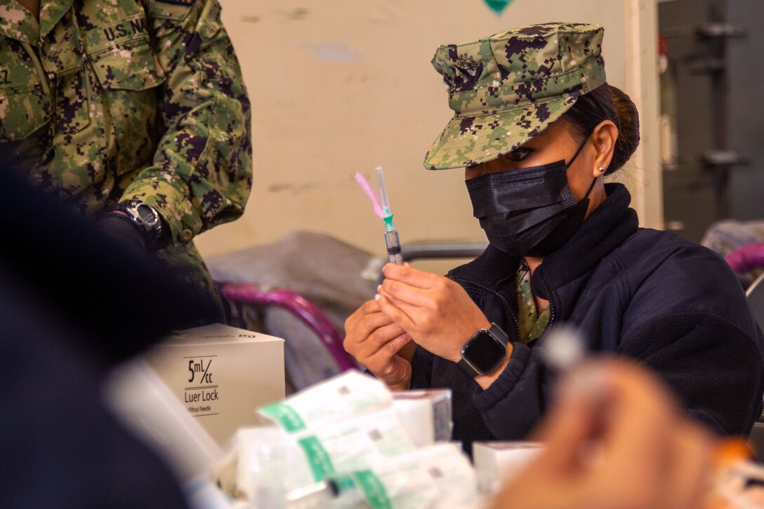 A sailor wearing a face mask holds a syringe while preparing it for a COVID-19 booster shot.