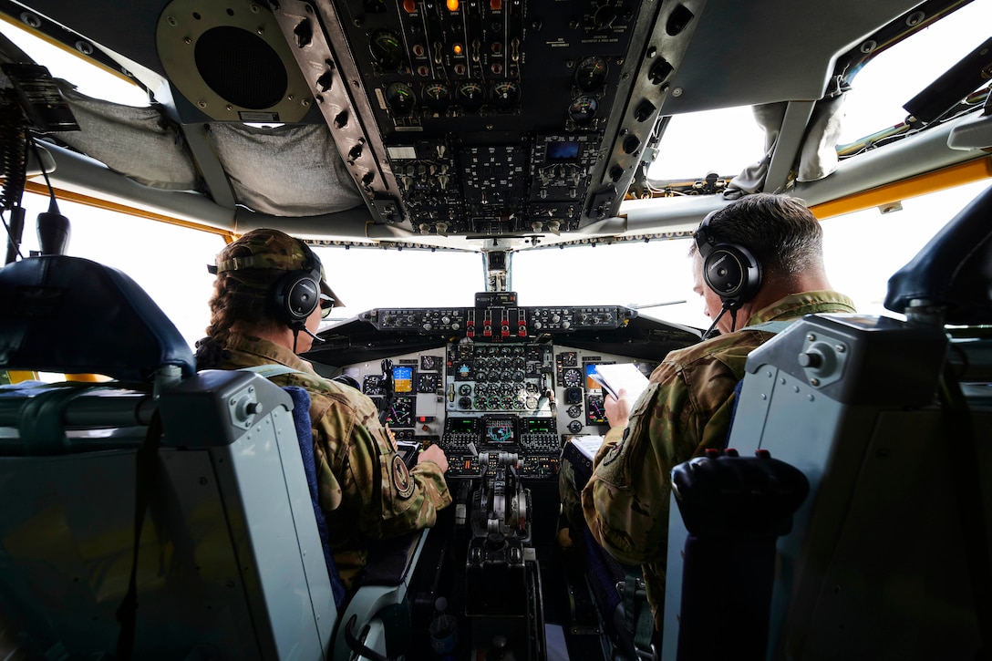 An airman seated in a cockpit pushes controls while another airman reviews paperwork.