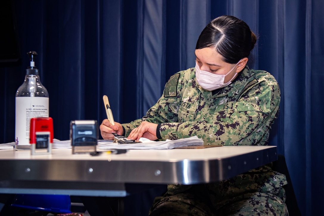 A sailor wearing a face mask sits at a desk tracking other sailors who have received a COVID-19 vaccine booster.