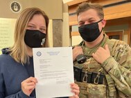 Staff Sgt. Alyssa Day, Air Force Mortuary Affairs Operations public affairs specialist, poses with her husband Staff Sgt. Spencer Day, 436th Security Forces Squadron, holding her Fiscal Year 2022 Senior Leader Enlisted Commissioning Program-Active Duty Scholarship recipient letter in the AFMAO atrium Dec. 25, 2021. AFMAO leadership coordinated with Day’s husband who was working Christmas day to surprise her with the notification of her selection. Day will attend Arizona State University, and upon finishing her degree will attend Officer Training School. (U.S. Air Force photo by Christin Michaud)