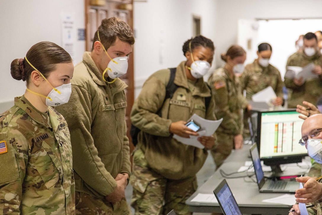 Guard members in OCP uniforms stand in a line before people in OCP uniforms at computers. They are going through a personnel deployment processing line and are in-processing for deployment throughout the state.