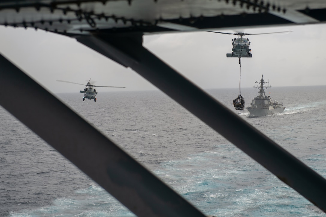 Two helicopters fly behind an aircraft carrier.