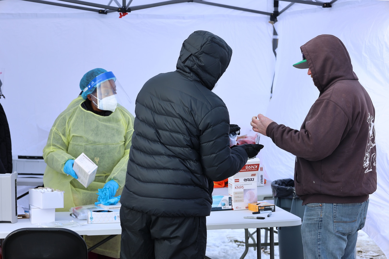 One care provider in PPE oversees two men engaged in COVID-19 test sampling under a tent.