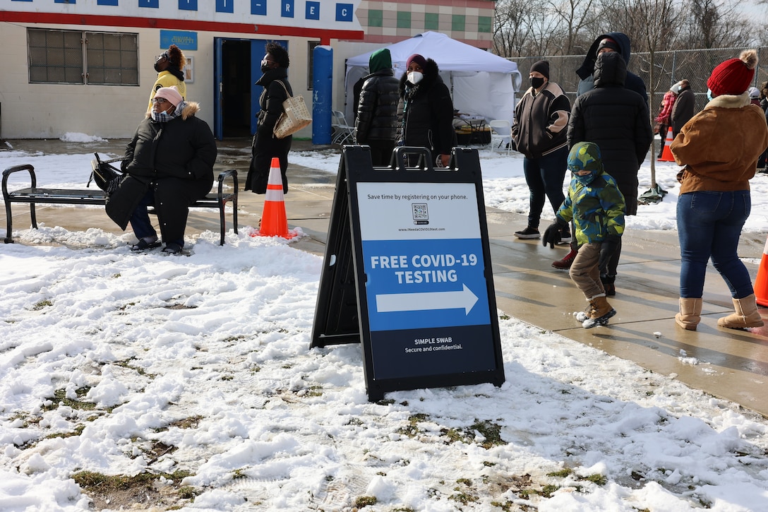 Citizens stand outside while snow is on the ground. A sign stating "Free COVID-19 testing" is on the ground at the center of the photo.