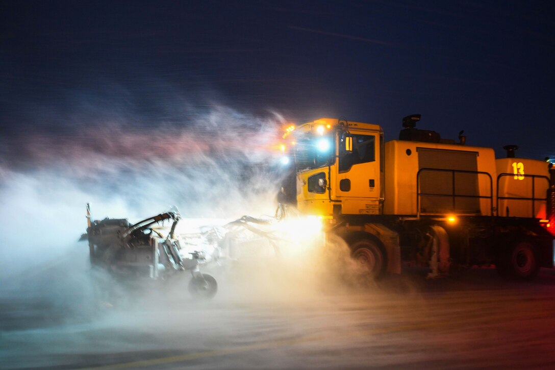 A snow plow and other snow removal vehicles remove snow at night using large headlights.