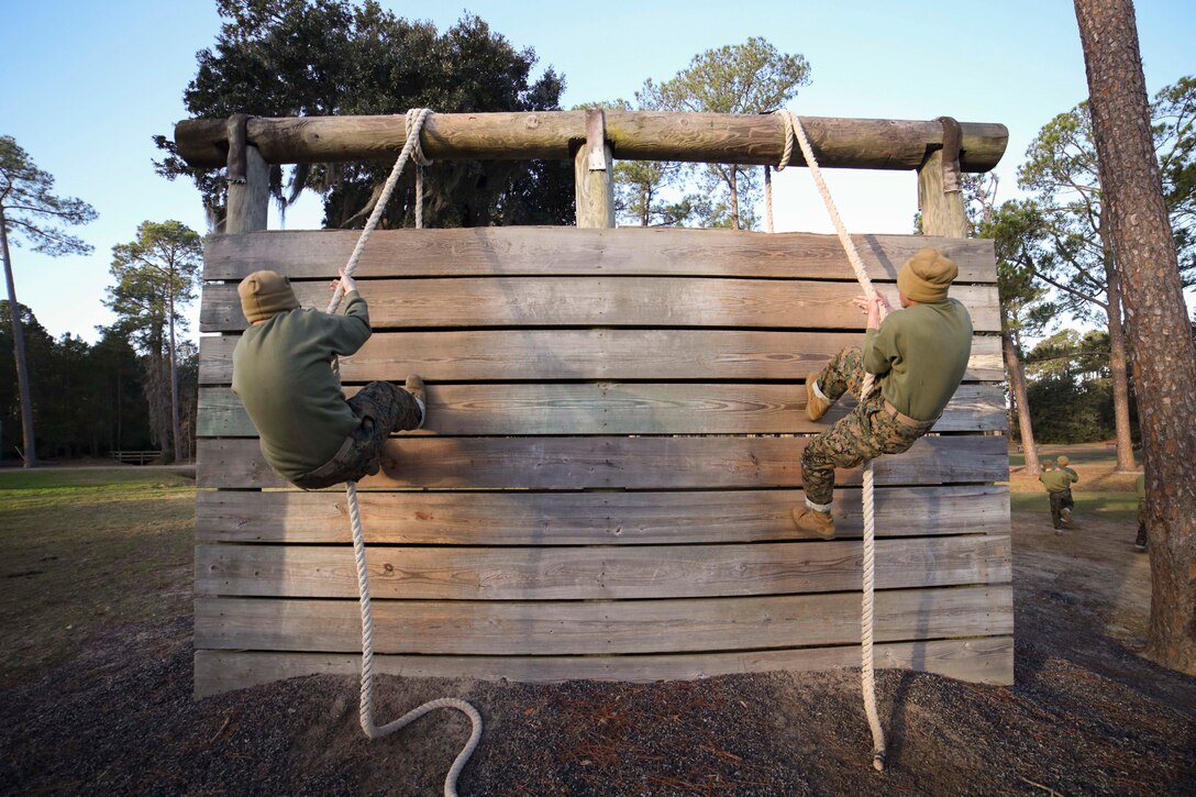 Two Marine Corps recruits climb a wooden wall with a rope.