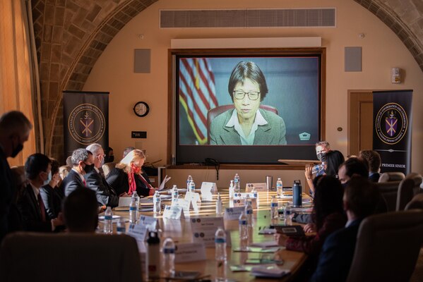 A woman on a large screen is meeting w=virtually with people around a large conference table.