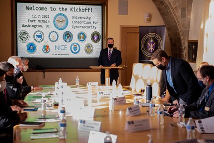 A man at a lectern speaks to a group at an adjacent table. The slide on the large screen behind him is welcoming the attendees.