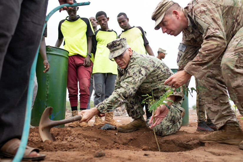 A service member shovels as another holds a sapling in a hole, as civilians stand and watch.