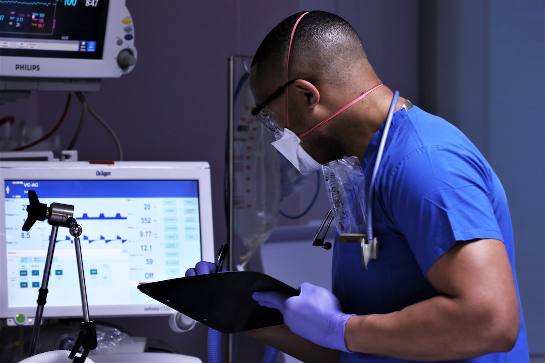 A sailor wearing a face mask and gloves holds a clipboard while looking at a screen.