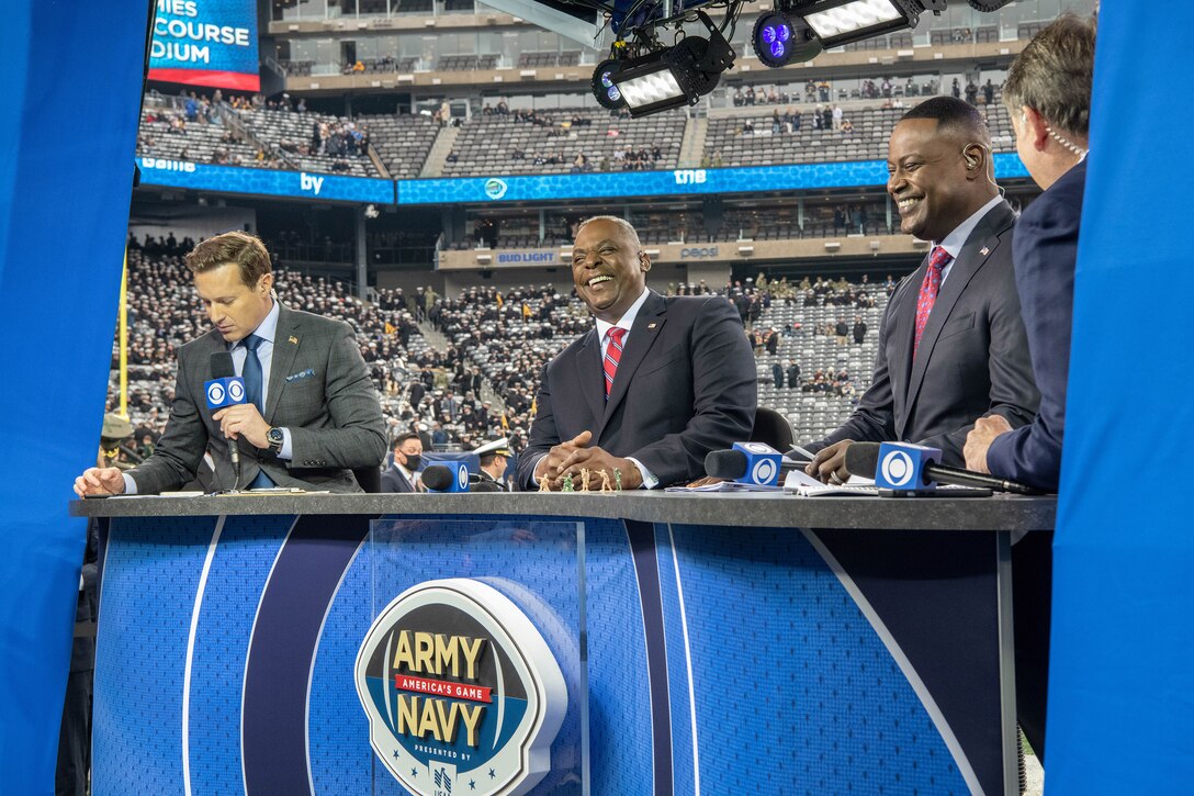 Secretary of Defense Lloyd J. Austin III laughs with others at a table in a crowded stadium.