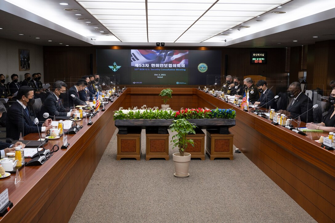 Officials talk at two long tables separated with a large screen on the wall at one end.