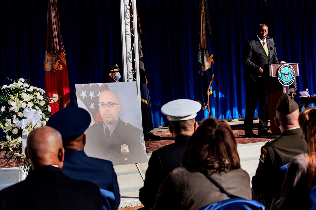 Secretary of Defense Lloyd J. Austin III speaks at a lectern next to large portrait photo on stage.