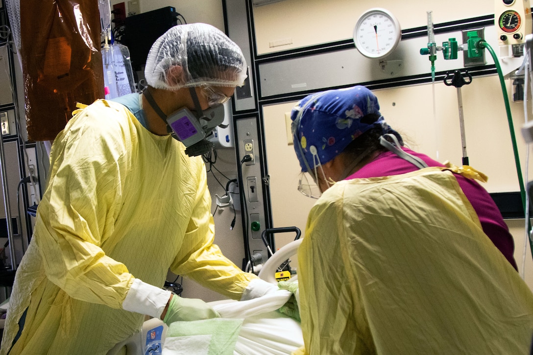 Two medical personnel in a hospital setting wearing personal protective equipment look down at something off camera.