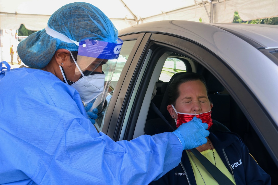 A soldier wearing personal protective equipment holds a nasal swab up to a woman sitting in a car.