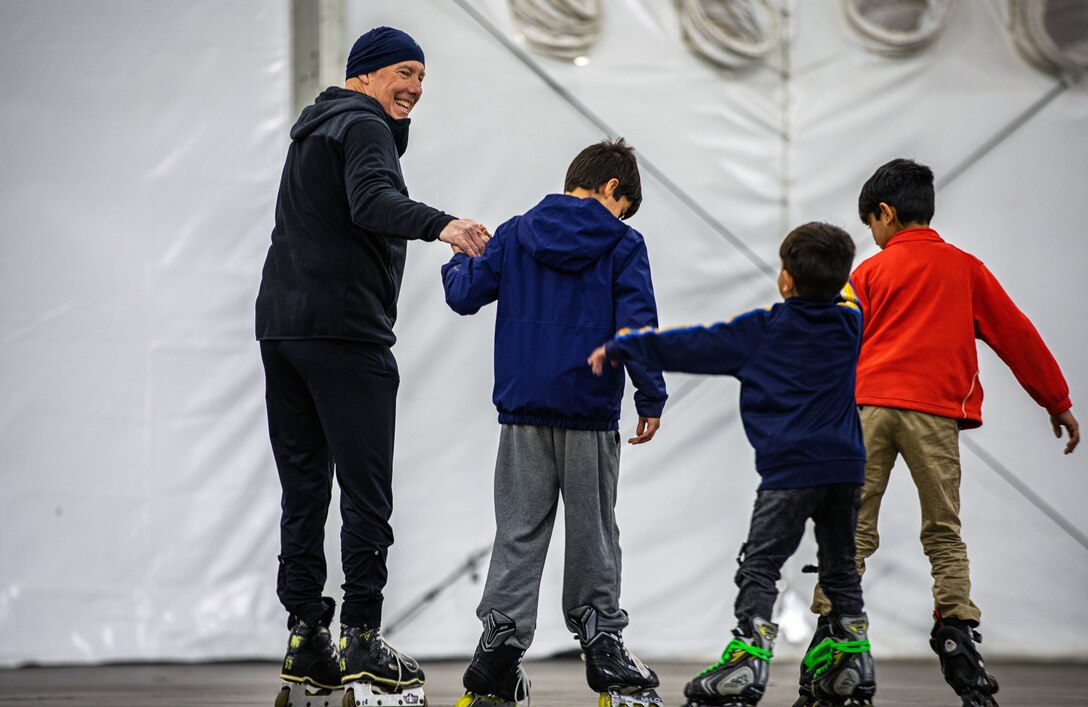 A man smiles and holds hands with a boy  as they roller-skate with two other boys.