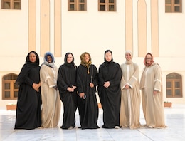 7 women dressed in black and tan robes and head coverings on a marble floor of a courtyard.