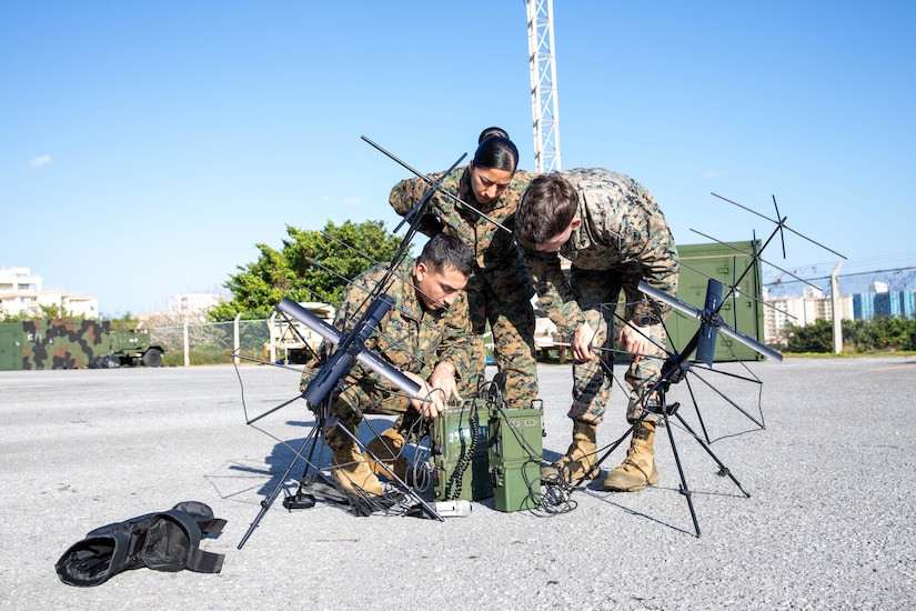 Three Marines work outside to adjust equipment on a paved surface.