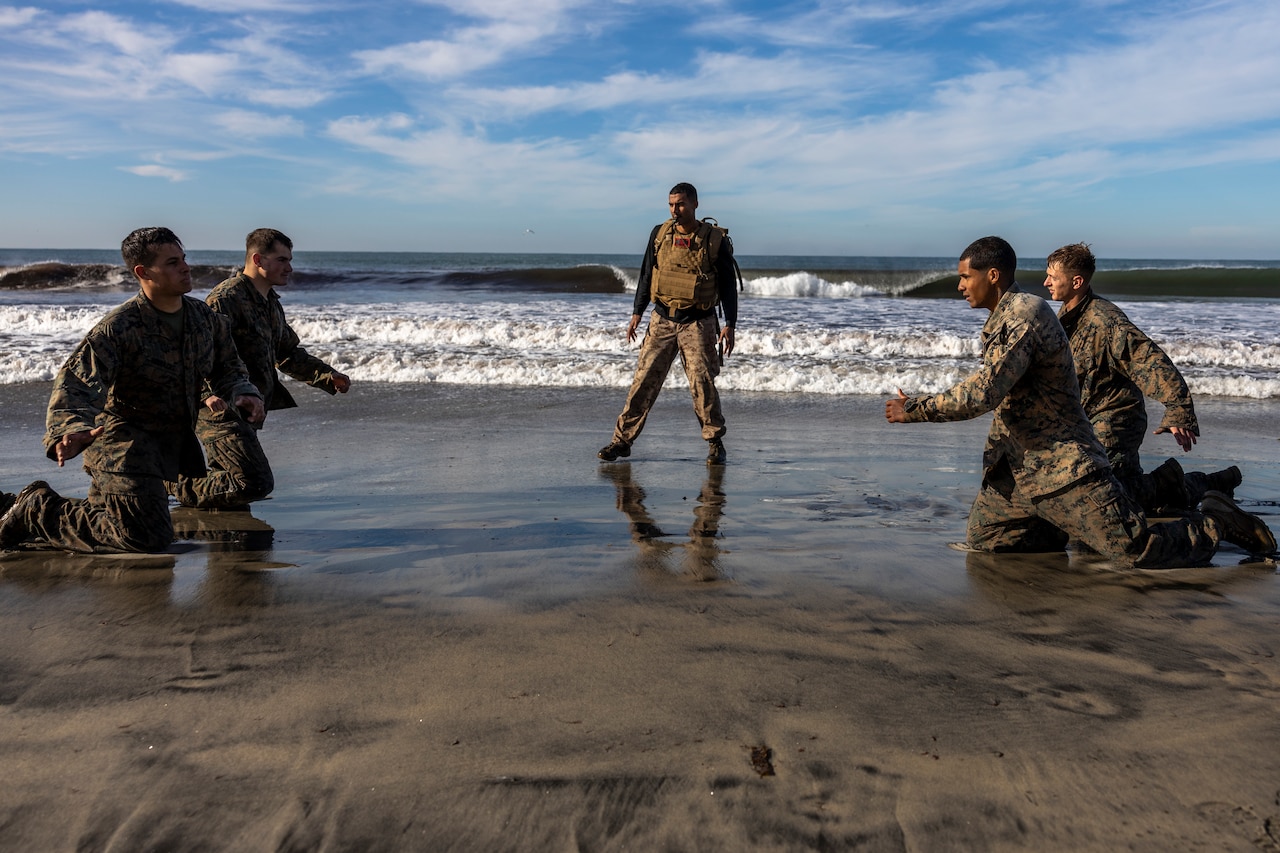 Two Marines kneel on a beach facing to other Marines; another Marines stands in between.
