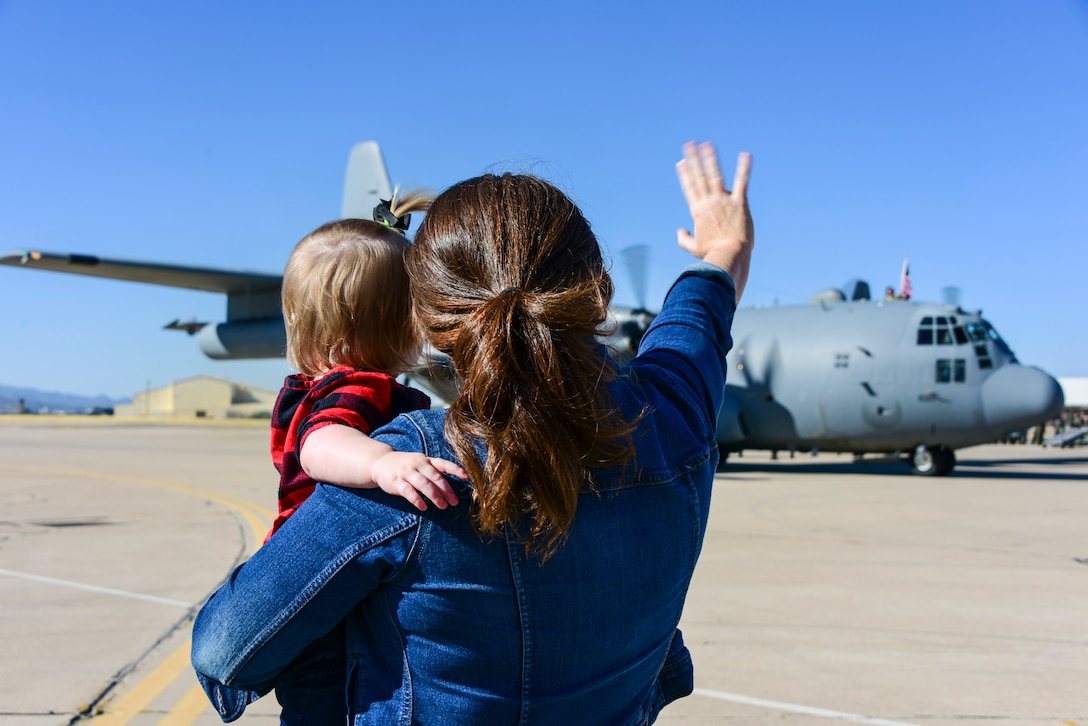 An adult holding a child waves toward an aircraft in the distance on a runway.