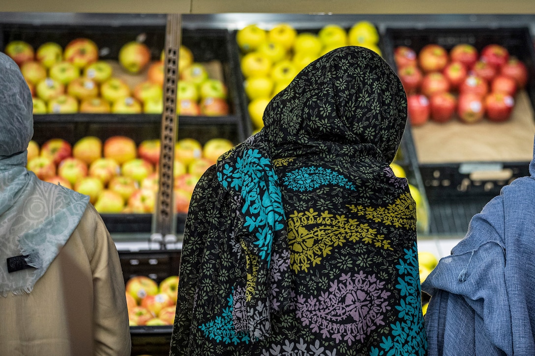 A person covered by a paisley hijab tilts their head and looks at bins of fruit in a grocery store.