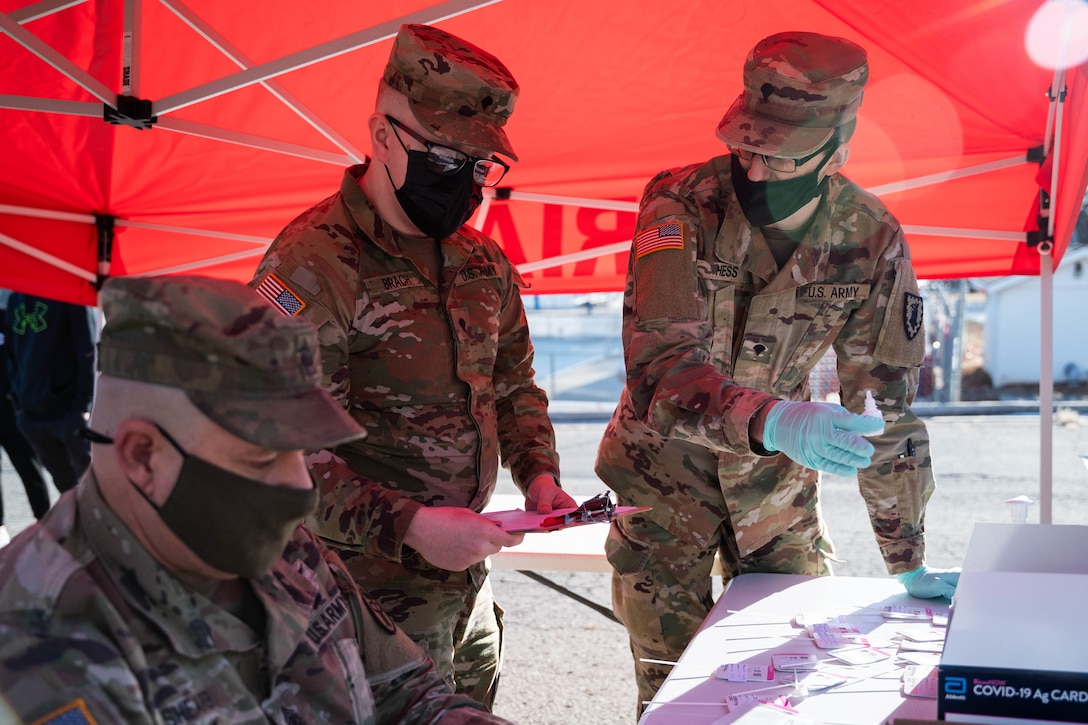 Three soldiers wear face masks and gloves; one holds a clipboard and another holds a small, plastic bottle.