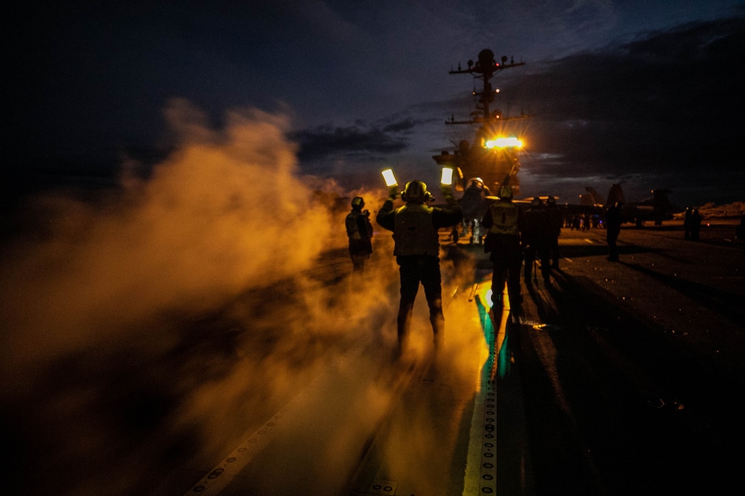 A sailor directs the pilot of a military aircraft on a ship at night.