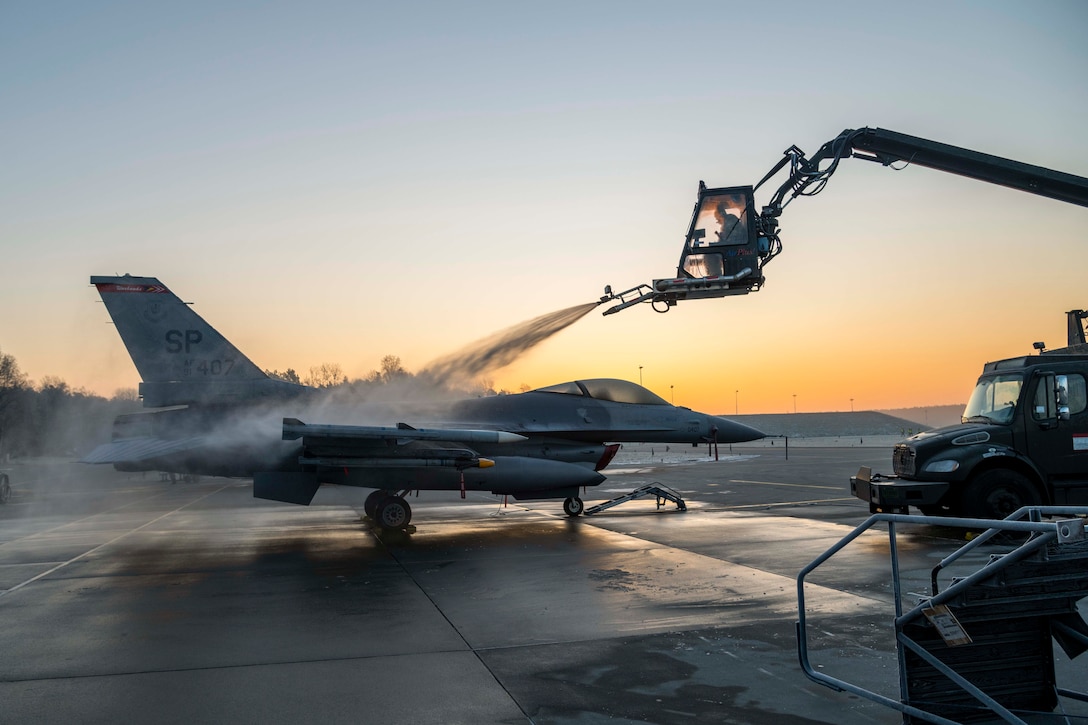 A crew member sitting in a crane sprays fluid onto an aircraft under a sunlit sky.